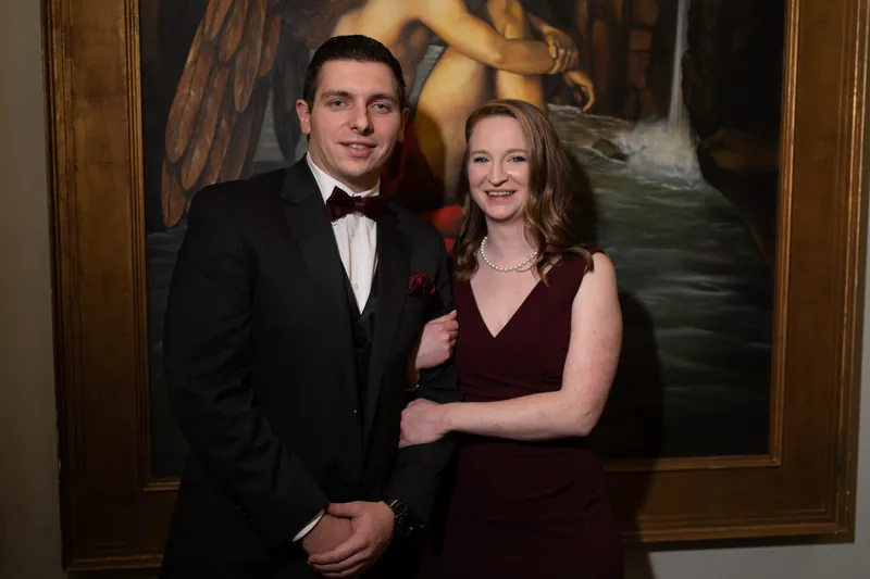 Michael and a guest pose together in formal attire in front of an ornate framed painting.