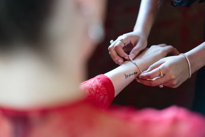 Hands tie a pink ribbon bow onto a white ribbon or sash during a getting-ready moment.