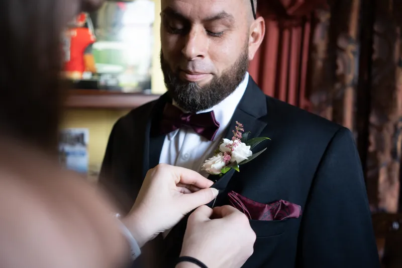 Michael looks down as someone adjusts a small floral boutonniere on his tuxedo lapel.