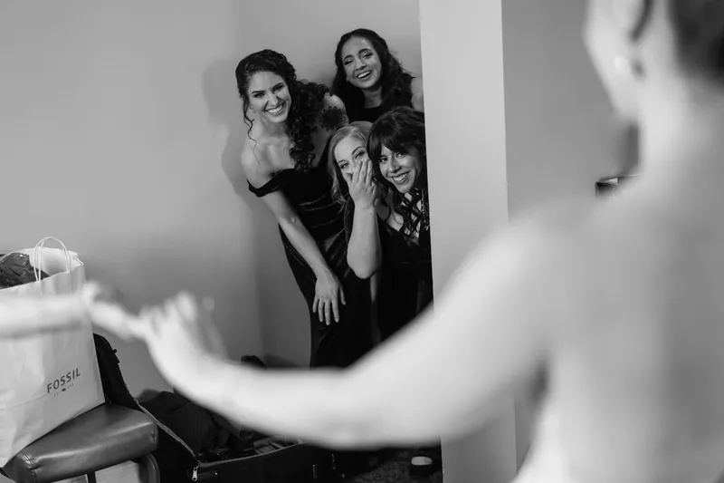 Simone laughs with two women in a mirror reflection during bridal preparations.