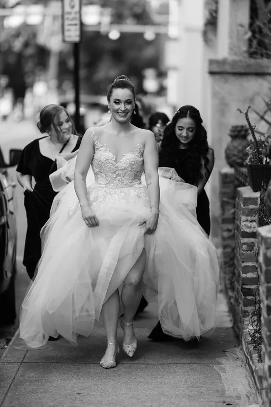 Simone walks through a courtyard in her beaded wedding dress, flanked by two women in black.