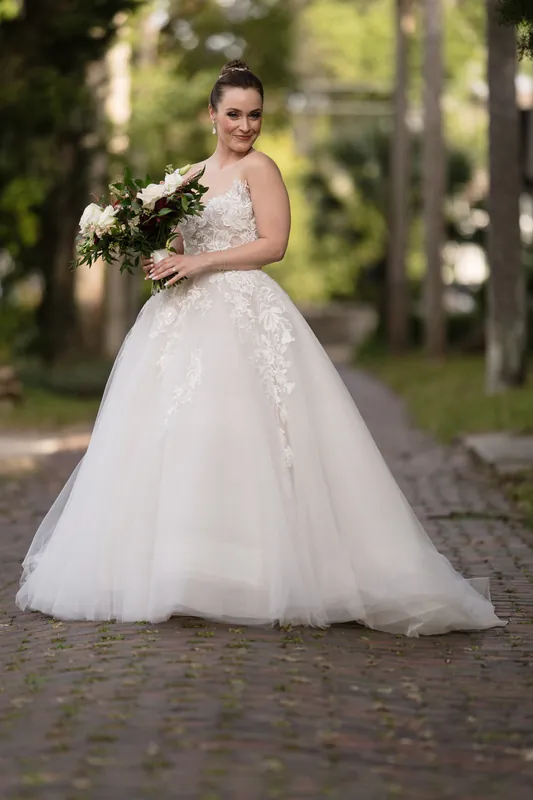 Simone wears a beaded lace ball gown and holds a cascading greenery bouquet on a brick pathway.