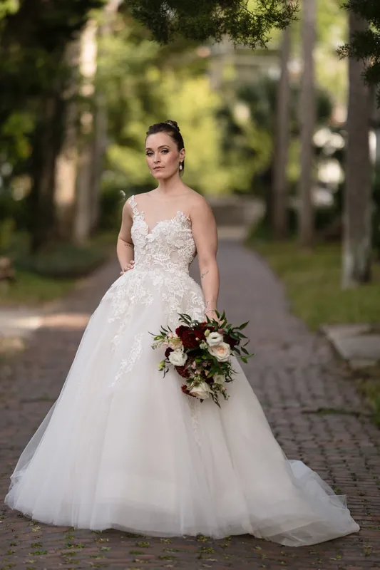 Simone stands in an embellished white gown holding a bouquet of white flowers and dark foliage.