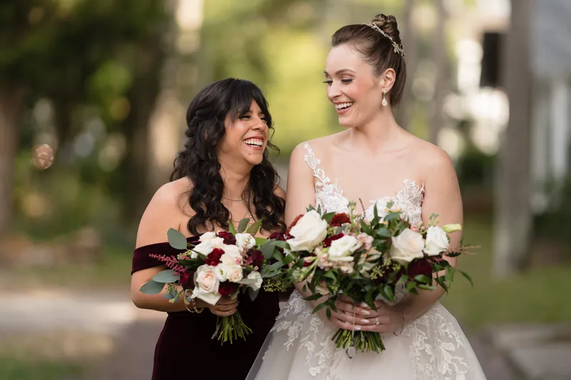 Simone and an attendant smile at each other while holding matching bouquets tied with burgundy ribbon.