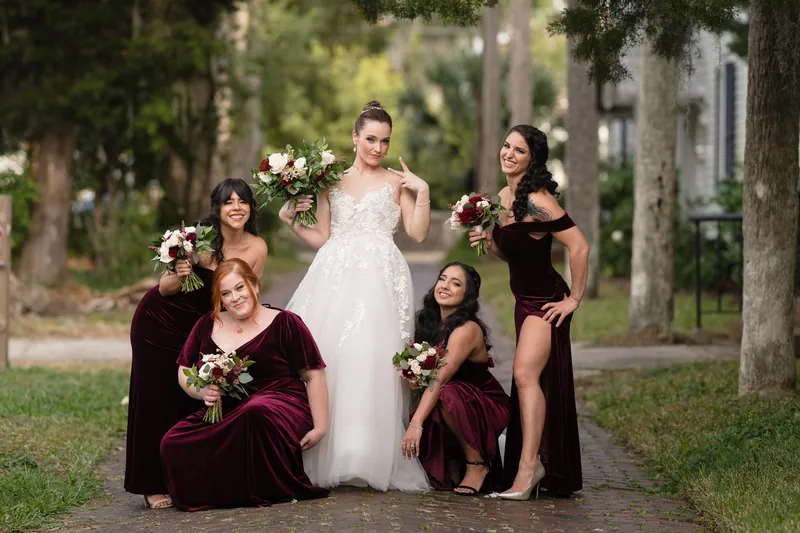Five attendants in burgundy velvet flank Simone on a tree-lined pathway as she holds her wedding bouquet.