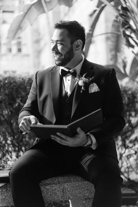 Michael reads from a booklet in black and white, seated outdoors in formal wedding attire.