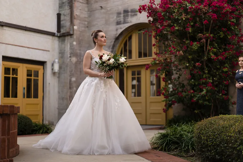Simone stands in a flowing white gown holding a cream-colored bouquet beneath ivy-covered arches.