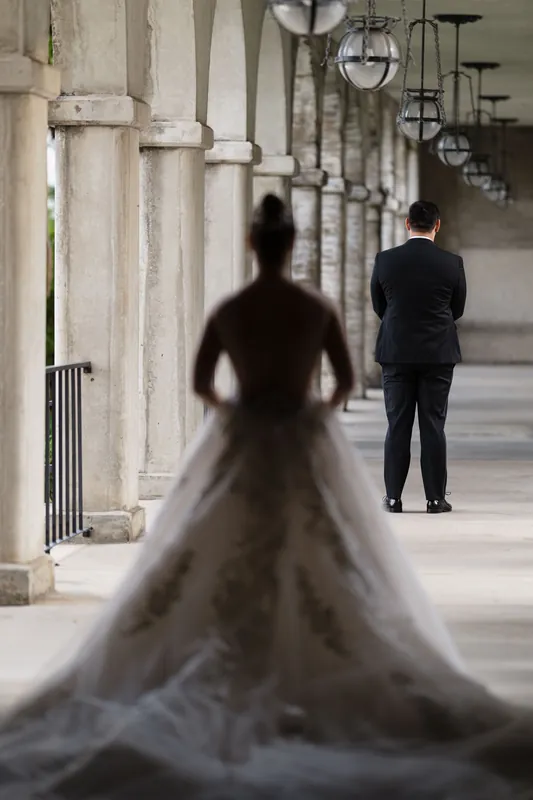 Simone walks toward Michael down a colonnade in her wedding dress as he waits in a dark suit.