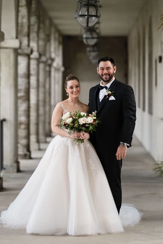 Simone and Michael walk together down a colonnade corridor, she carrying a white and green bouquet, he wearing a boutonniere.