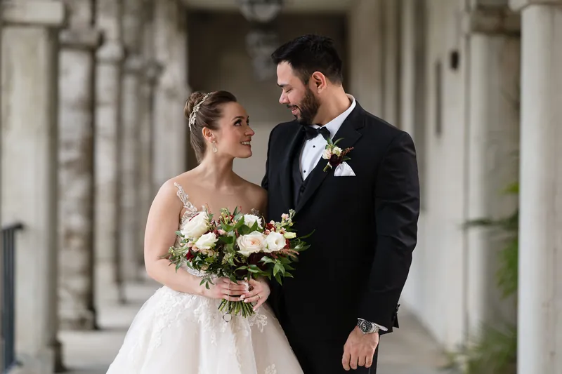 Simone looks toward Michael as they stand beneath arched columns, both smiling with her bouquet between them.