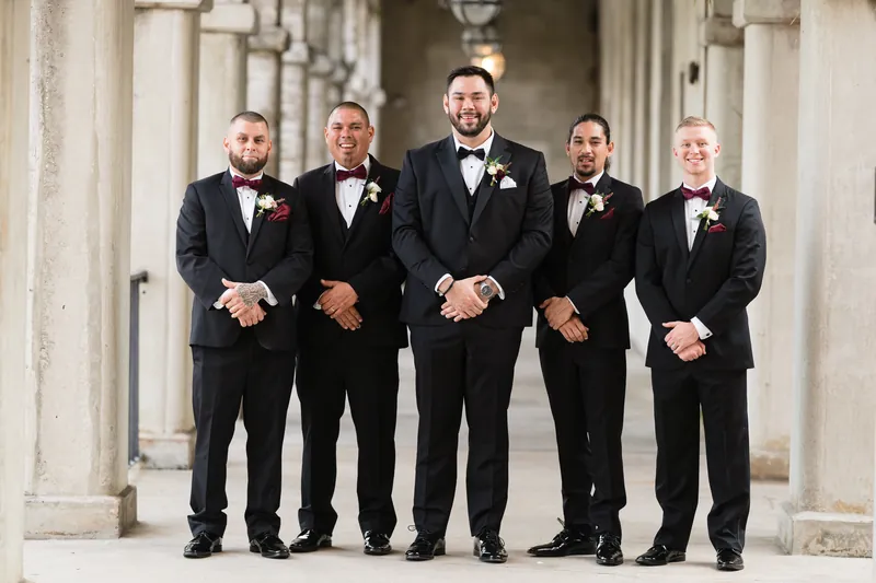 Five groomsmen in black tuxedos with burgundy boutonnieres stand shoulder to shoulder on the resort's covered veranda.