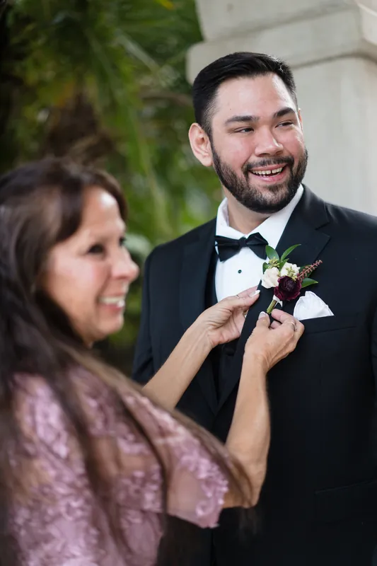 A woman adjusts Michael's burgundy and cream boutonniere as he smiles at Casa Monica Resort & Spa.