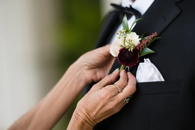 Simone's hand holds a deep burgundy rose boutonniere against Michael's black tuxedo jacket at Casa Monica Resort & Spa.