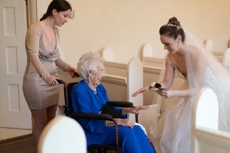 Two attendants help an elderly woman in a blue dress sit while Simone adjusts her corsage indoors.