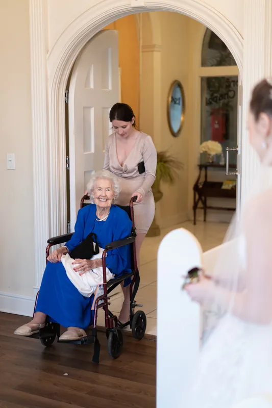 An elderly woman in blue sits in the foyer while Simone leans down to greet her beneath an arched doorway.