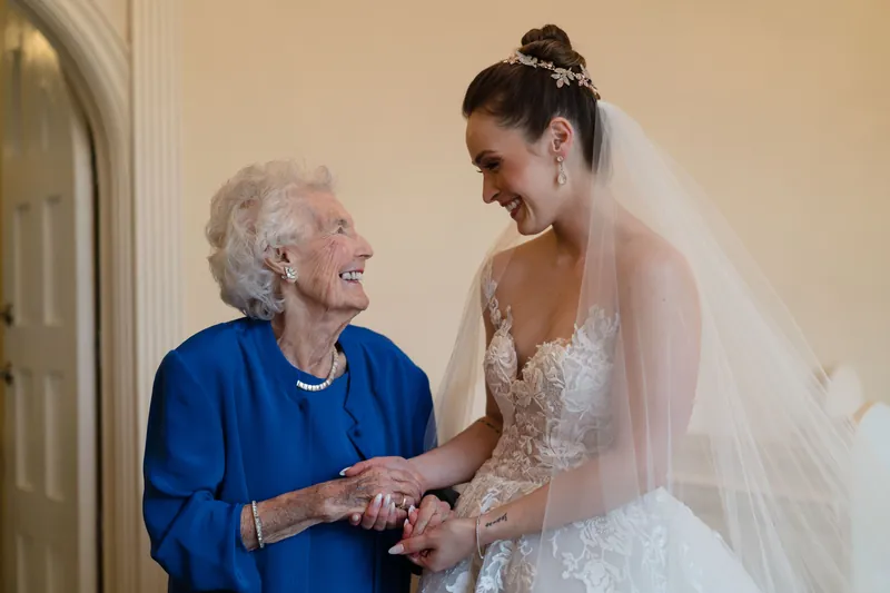 Simone in her beaded wedding dress smiles while holding hands with an older woman wearing a blue dress and pearl necklace.