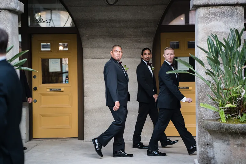 Three men in dark suits walk through a covered stone archway with yellow wooden doors flanking the passage.