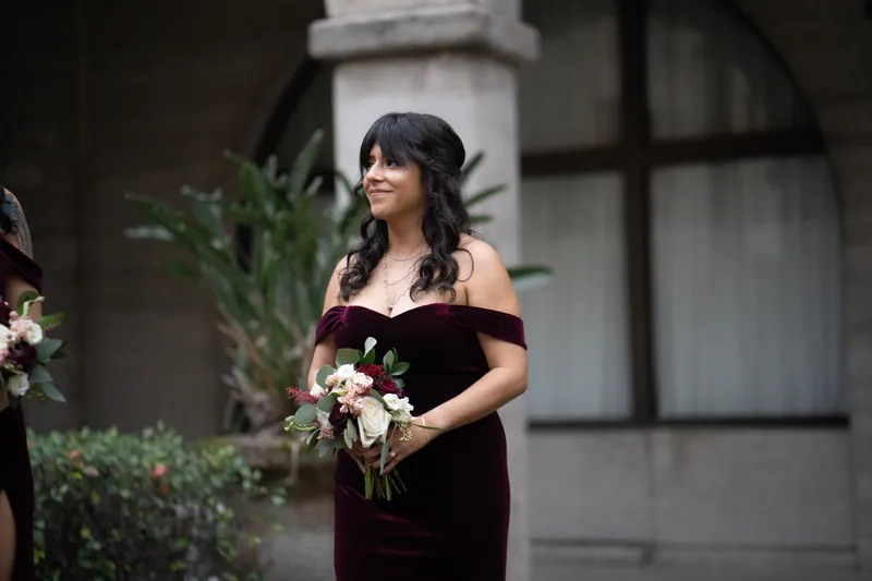 Simone holds her bouquet while wearing an off-shoulder burgundy velvet gown in the courtyard.