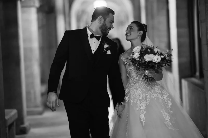Michael and Simone walk together down a colonnade in black and white, Simone holding a white floral bouquet.