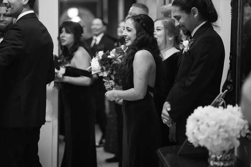Simone smiles radiantly surrounded by formally dressed guests in a hallway, holding her white bouquet.