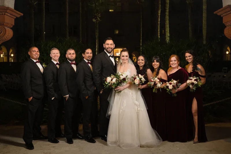 Simone in a white gown holds her bouquet while standing with Michael and their wedding party under palm trees at night.