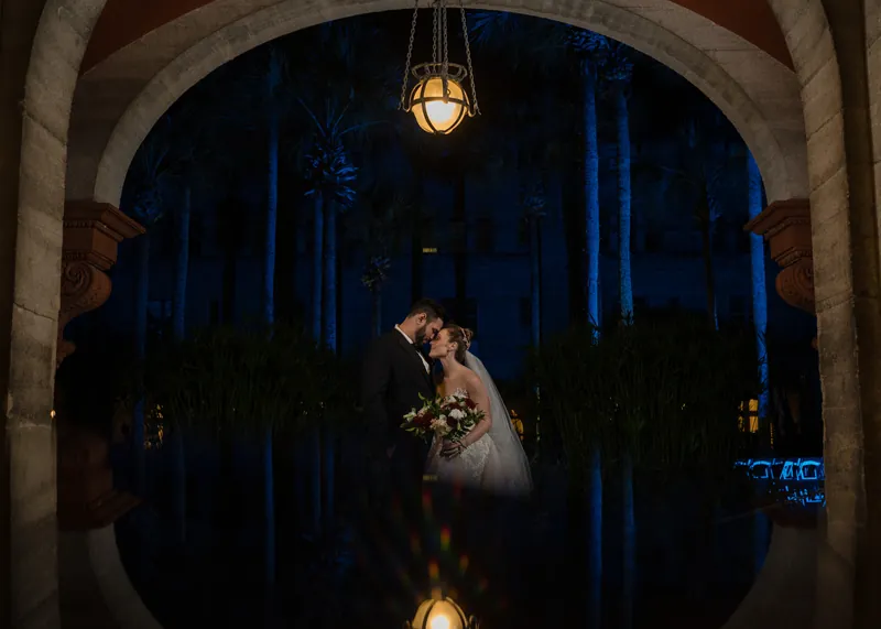Simone and Michael share a kiss framed by an arched stone doorway with blue-lit palms visible beyond.