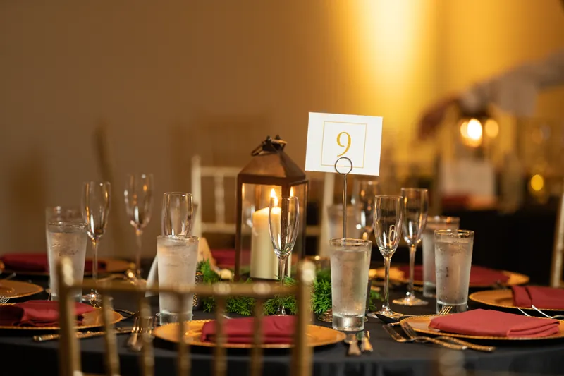Table setting features a numbered card, water glasses, and greenery accents on burgundy linens.