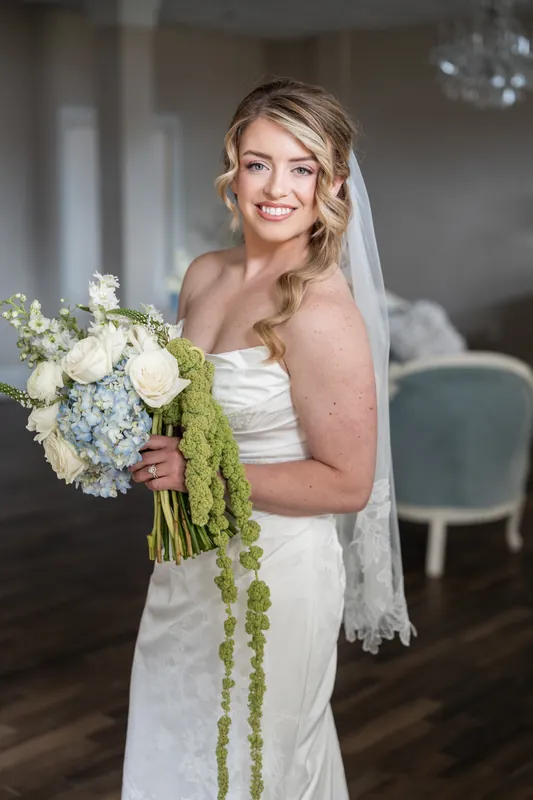 Skye smiles while holding a white and green bouquet in her strapless wedding dress and veil at The White Room.