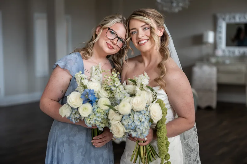 Skye's mother hugs her from behind while holding a large white and blue bridal bouquet indoors.