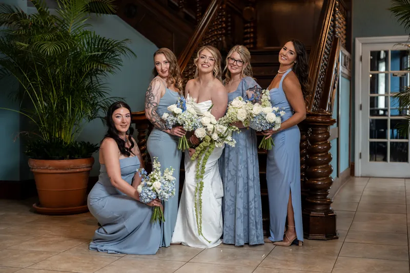 Five bridesmaids in varying shades of blue dresses hold white and green bouquets on The White Room's tiled floor.