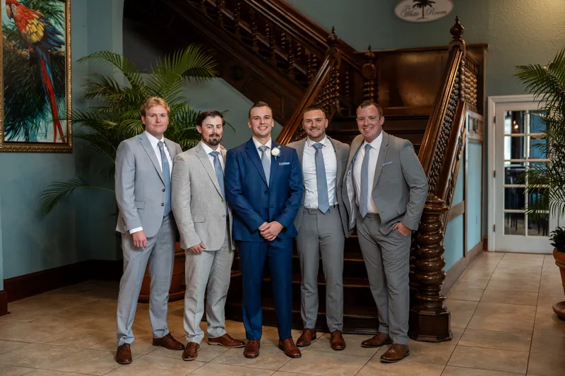 Five groomsmen in gray and navy suits stand together in front of a wooden staircase at The White Room.