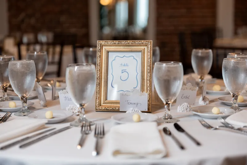 A gold-framed table number five sits among white place settings and glassware at a reception table.