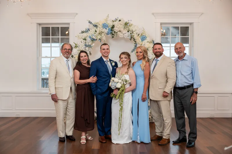 Skye and Elliot stand with seven family members beneath a blue and white floral arch at The White Room.