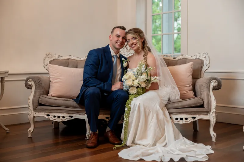 Elliot and Skye sit together on a cream-colored sofa, holding her bouquet and smiling at each other.