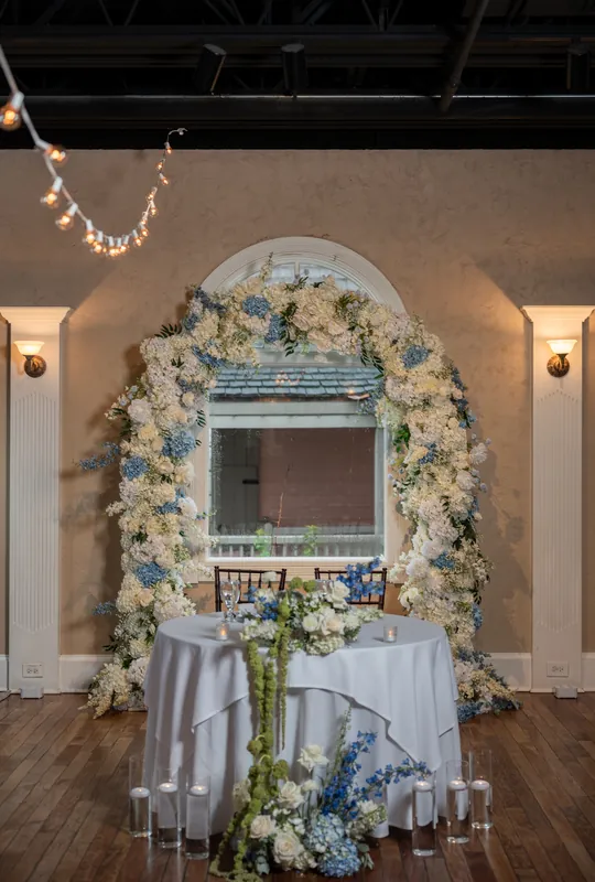 A sweetheart table decorated with white and blue florals frames a mirrored arch at The White Room.