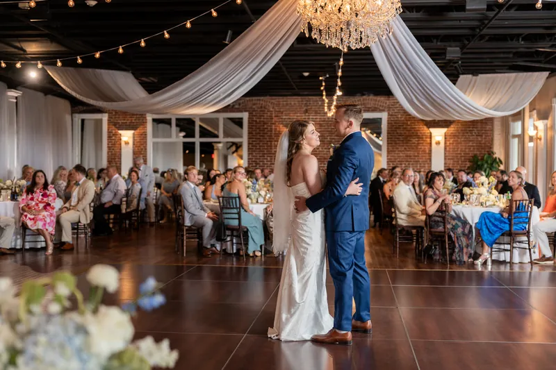 Elliot in a blue suit and Skye in a white gown dance at the center of the White Room's ballroom beneath a crystal chandelier and draped fabric.