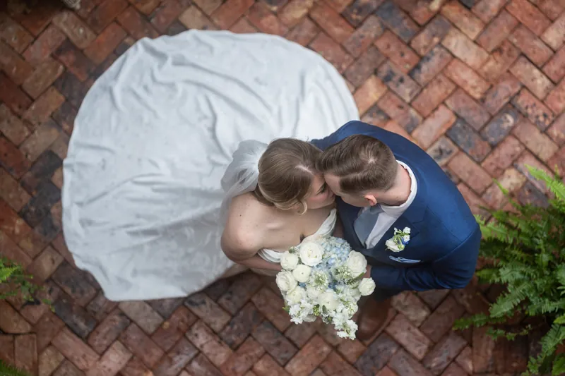 Overhead view of Skye and Elliot kissing on a herringbone brick floor, her white gown and his blue suit framing their white bouquet.