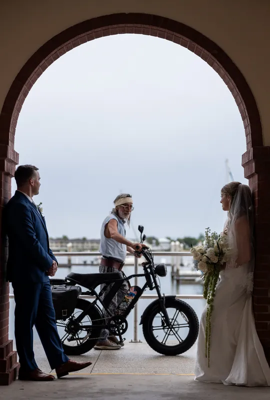 Skye and Elliot stand beneath the White Room's arched entrance with a guest on a vintage bicycle by the water.