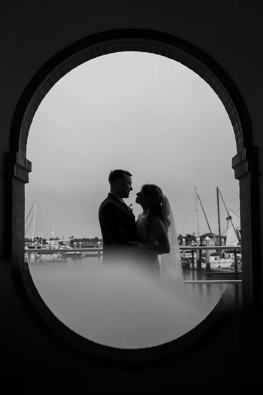 Black and white portrait of Skye and Elliot kissing, framed by an oval window with marina views behind them.