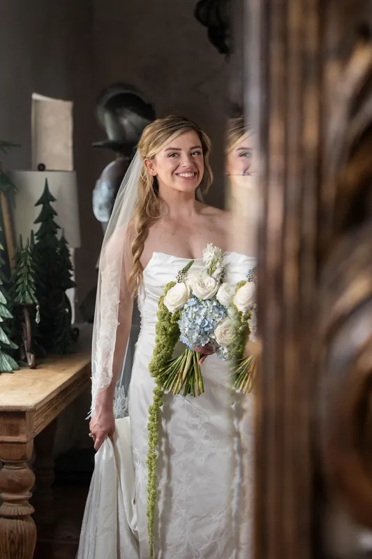 Skye holds her white bouquet and adjusts her veil indoors, sunlight streaming across her face at the White Room.