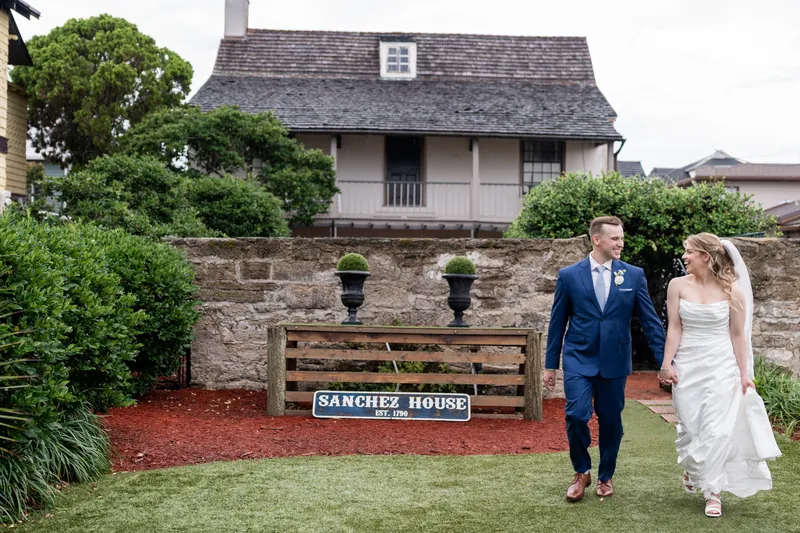 Elliot in navy suit and Skye in white wedding dress walk hand in hand past the Sanchez House at White Room.