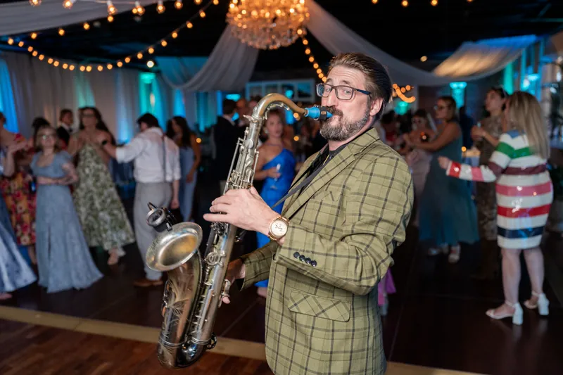 Saxophonist in a yellow plaid jacket performs during the reception at White Room with dancing guests behind him.