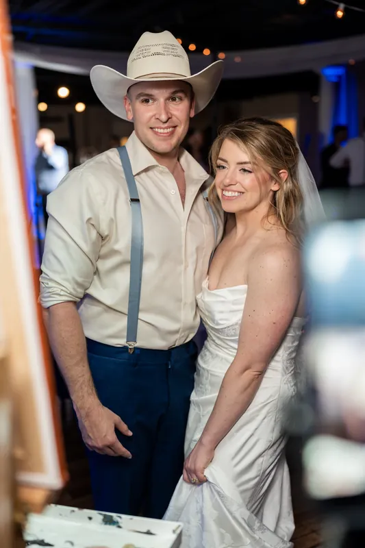 Skye and Elliot stand together smiling, with Elliot wearing a cowboy hat and blue jeans at the White Room reception.
