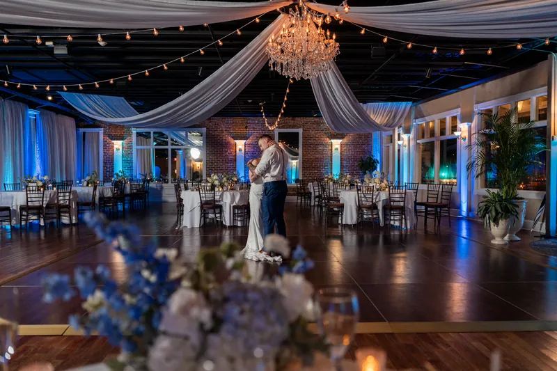 Skye and Elliot dance under draped fabric and a crystal chandelier in the White Room's ballroom with blue uplighting.