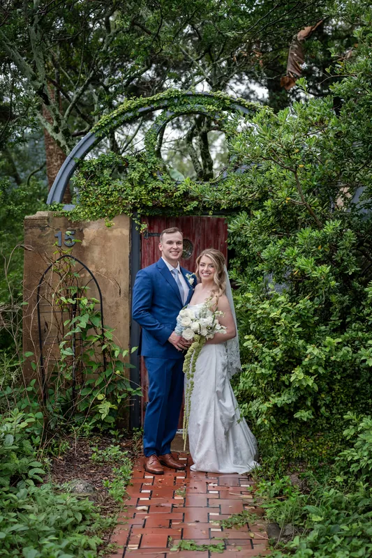 Skye in a white gown and Elliot in navy stand together under a ivy-covered archway on a brick garden path.