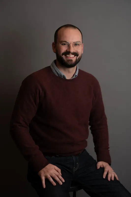 Man in a maroon sweater and dark jeans smiles at the camera against a neutral gray backdrop in a St. Augustine studio.