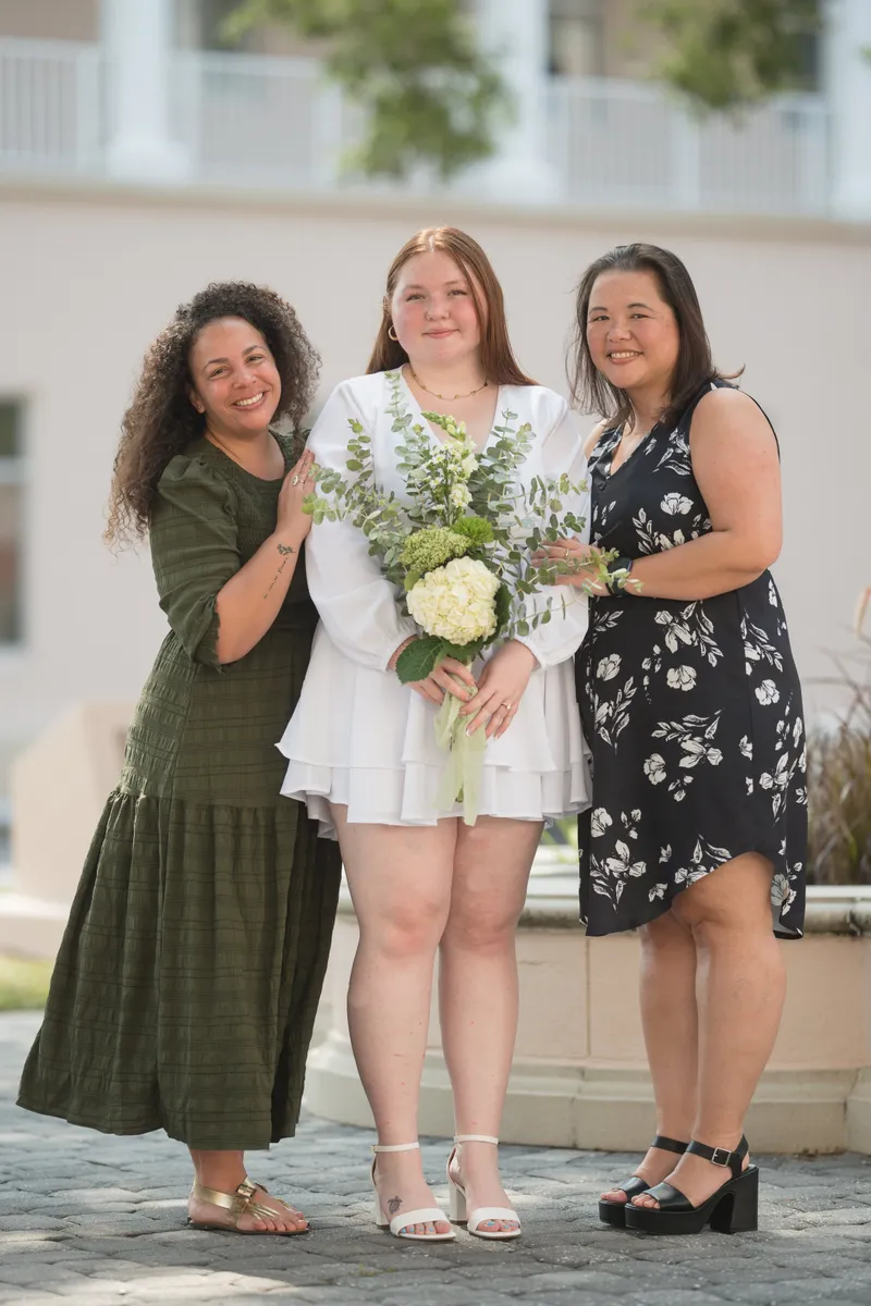 A joyful bride in white holds her bouquet while celebrating with two close friends or family members after her courthouse elopement ceremony.