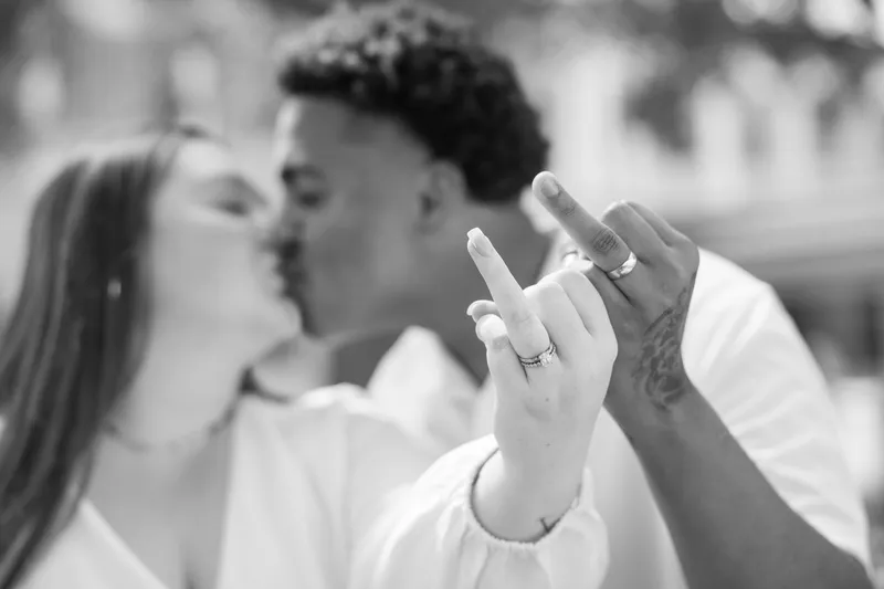 A tender black and white moment of newlyweds showing off their wedding rings with an intimate kiss in the background.