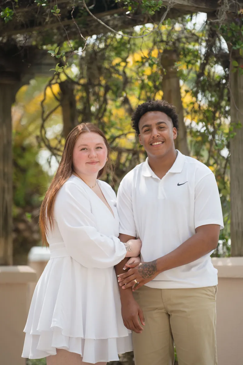 A happy couple poses together outdoors in coordinating white outfits, with the bride in a flowing white dress and groom in a white polo shirt.