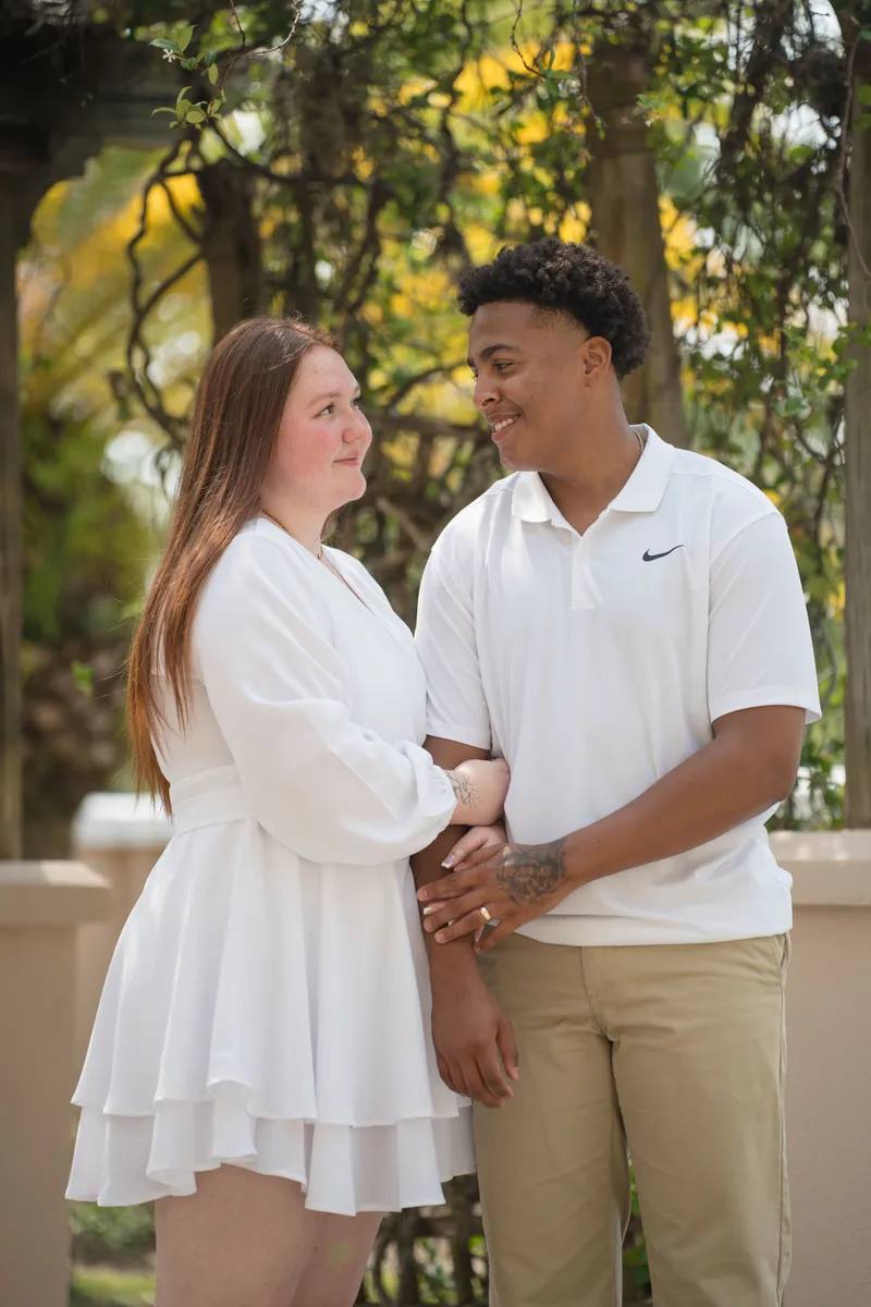 A couple shares an intimate moment looking into each other's eyes while embracing in matching white outfits under beautiful tree foliage.